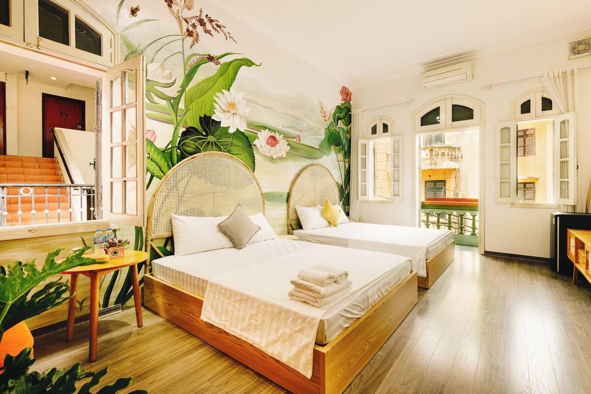 Een hotelkamer met twee bedden, een houten vloer, een groot raam en een muurschildering van bloemen en planten.