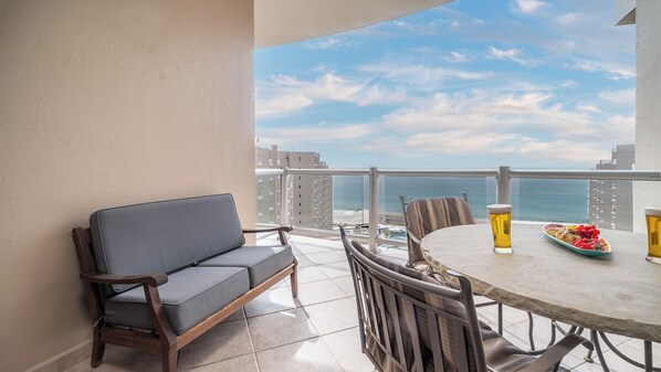 Uma varanda com sofá, mesa de jantar, cadeiras e vista para o oceano e edifícios.