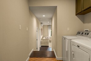 A laundry room with a washing machine and dryer, a wooden floor, and a hallway leading to a bedroom with a bed and curtains.
