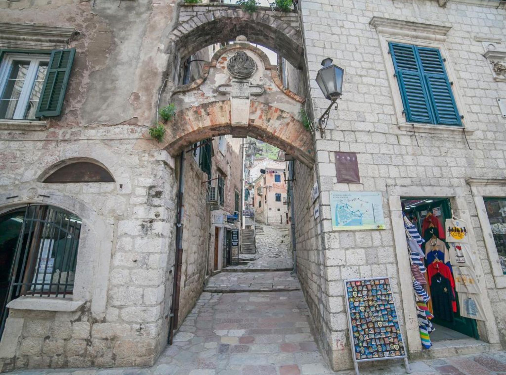 Una calle estrecha con un arco de piedra, tiendas a ambos lados y una pequeña exhibición de recuerdos frente a una tienda.