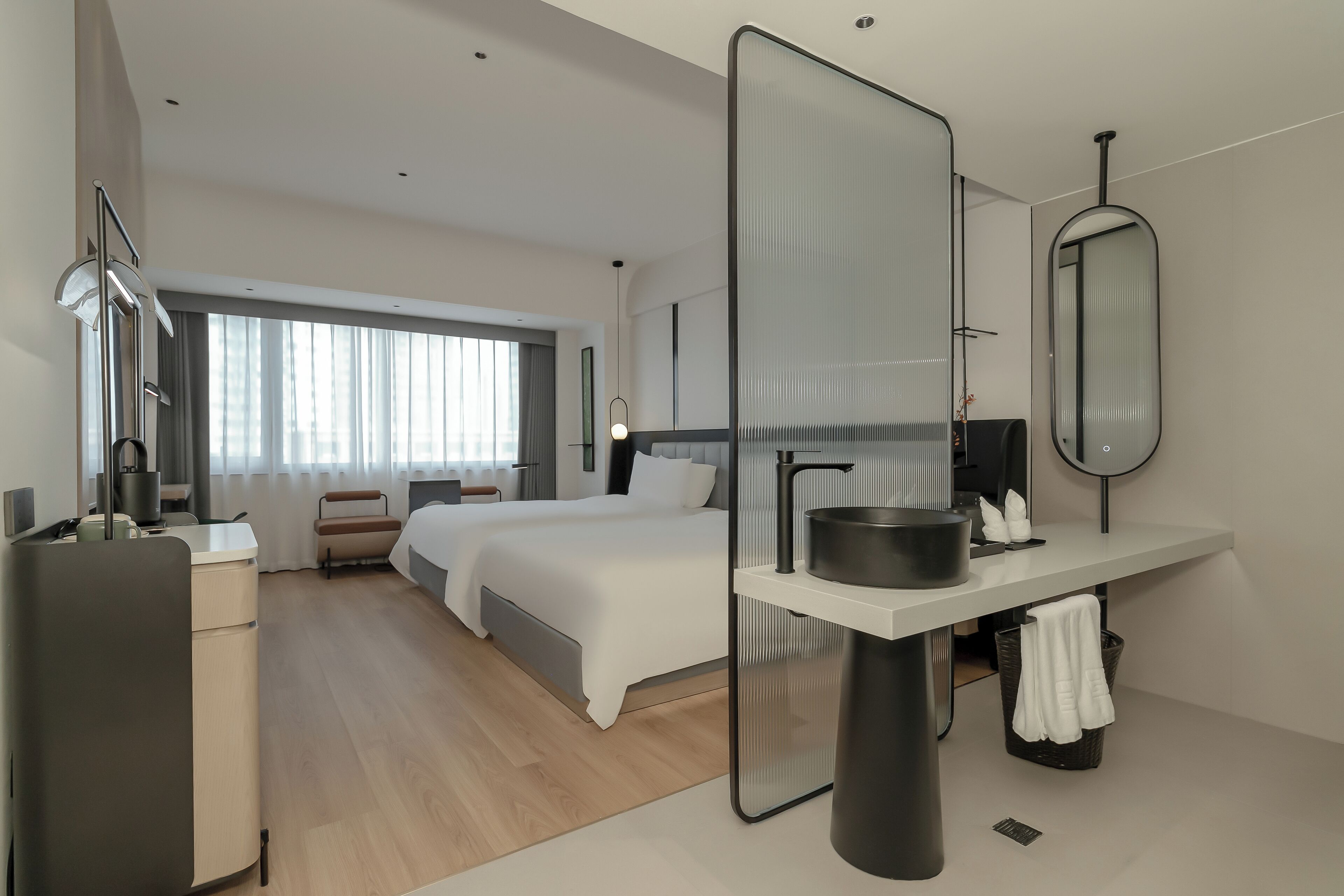 A modern hotel room with a large bed, a desk with a computer, a mirror, and a window with curtains.