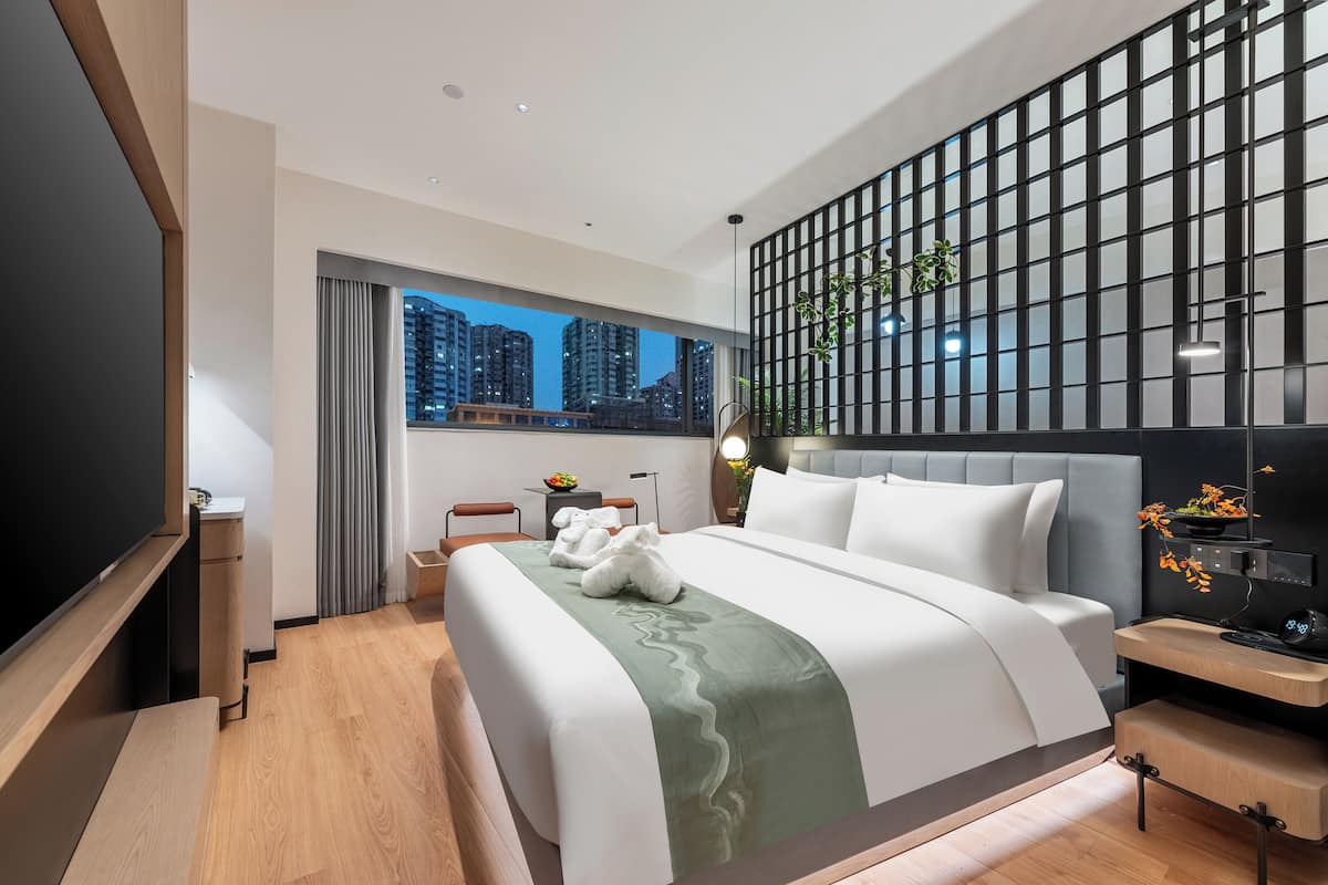 A modern hotel room with a large bed, a TV, a desk with a lamp, and a city view.