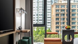 A modern hotel room with a large window, a flat-screen TV, a green chair, a wooden side table with a fruit bowl, and a round side table. - K.Space KaiShi hotel (Guangzhou)