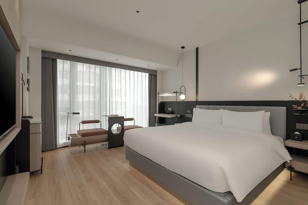A modern hotel room with a large bed, a desk, a chair, a TV, and a large window with curtains.