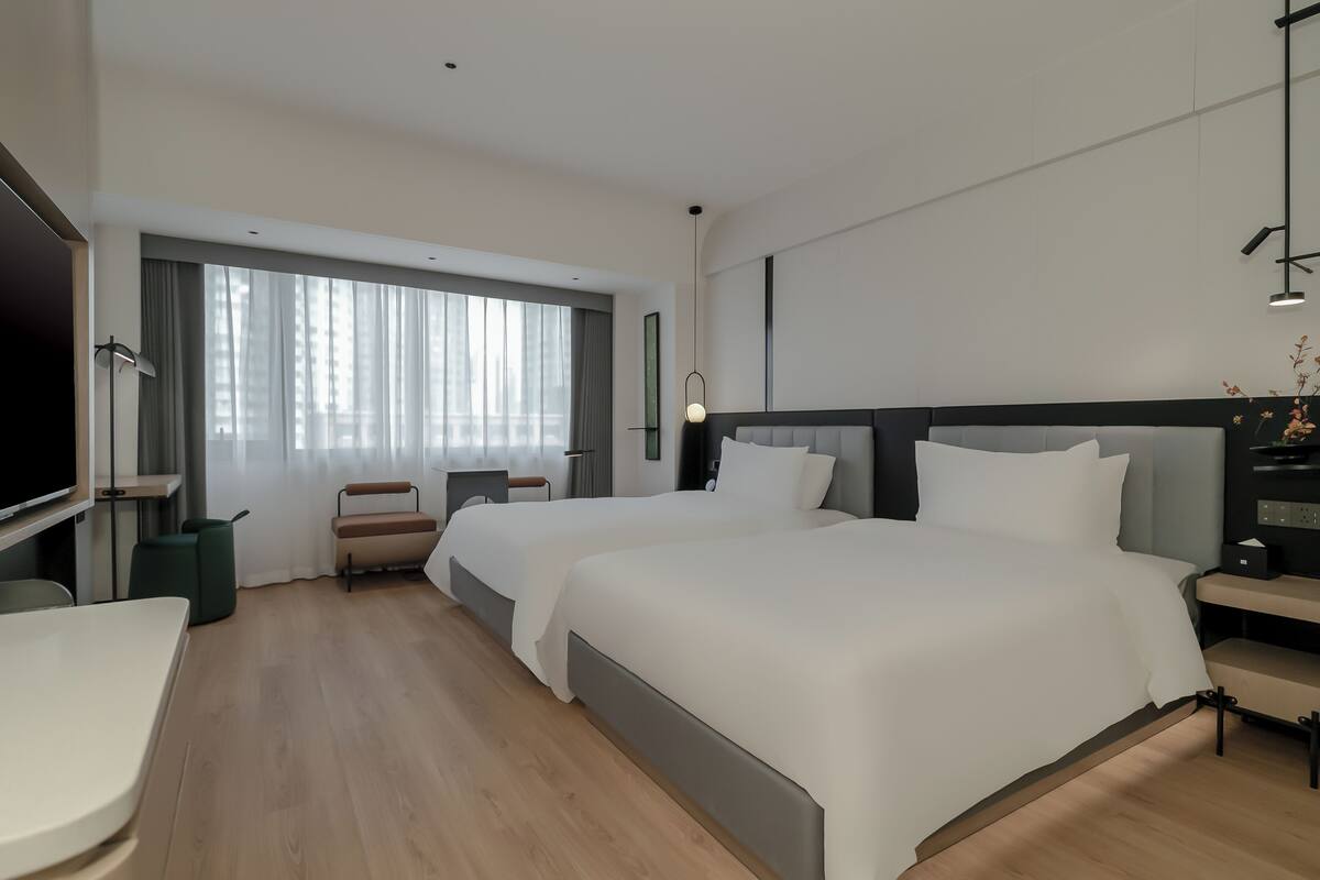 A modern hotel room with two beds, a flat-screen TV, a desk, and a large window with curtains.
