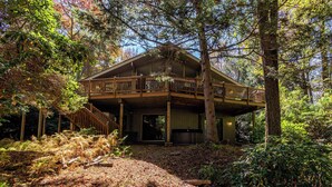 A two-story wooden cabin with a balcony, surrounded by dense forest. - Hidden Treasure Chalet Hot Tub | Game Room (Pocono Pines)