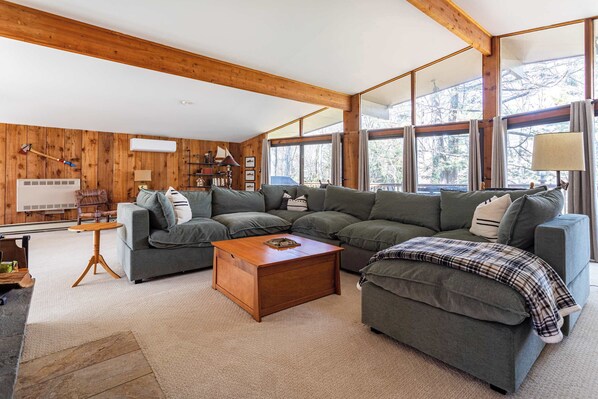 A cozy living room with a sectional sofa, a wooden coffee table, and large windows offering a view of trees. - Hidden Treasure Chalet Hot Tub | Game Room (Pocono Pines)