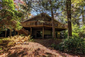 A two-story wooden cabin with a balcony, surrounded by dense forest.