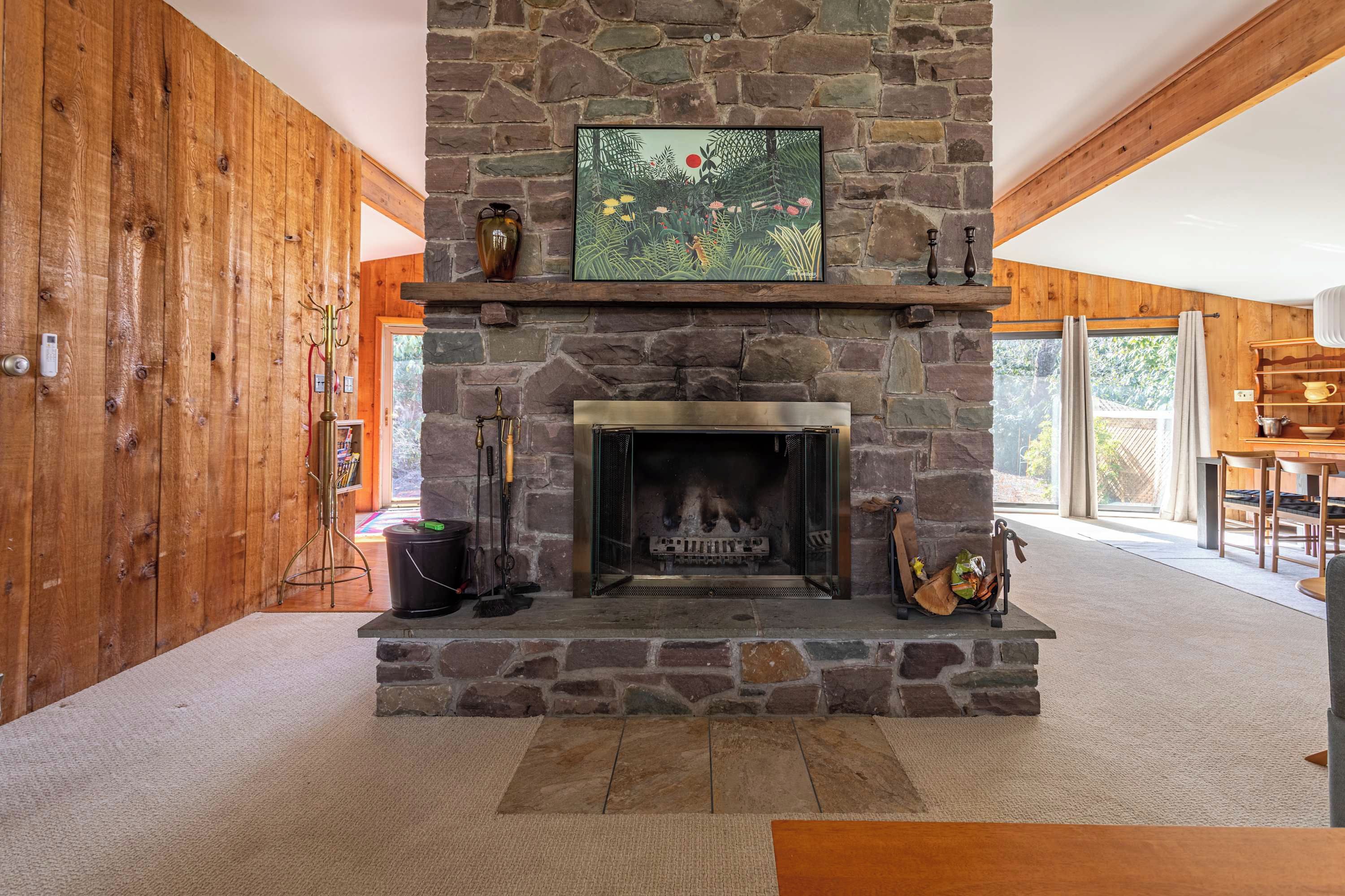 A stone fireplace with a framed picture above it, a wooden wall, and a view of the outdoors through a glass door.