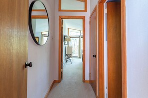 A hallway with a wooden door, a mirror, and a view into a room with a window and a chair. - Hidden Treasure Chalet Hot Tub | Game Room (Pocono Pines)