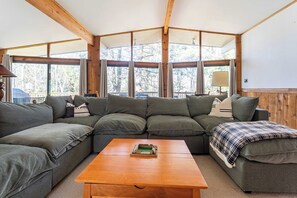 A cozy living room with a sectional sofa, a wooden coffee table, and large windows offering a view of trees.