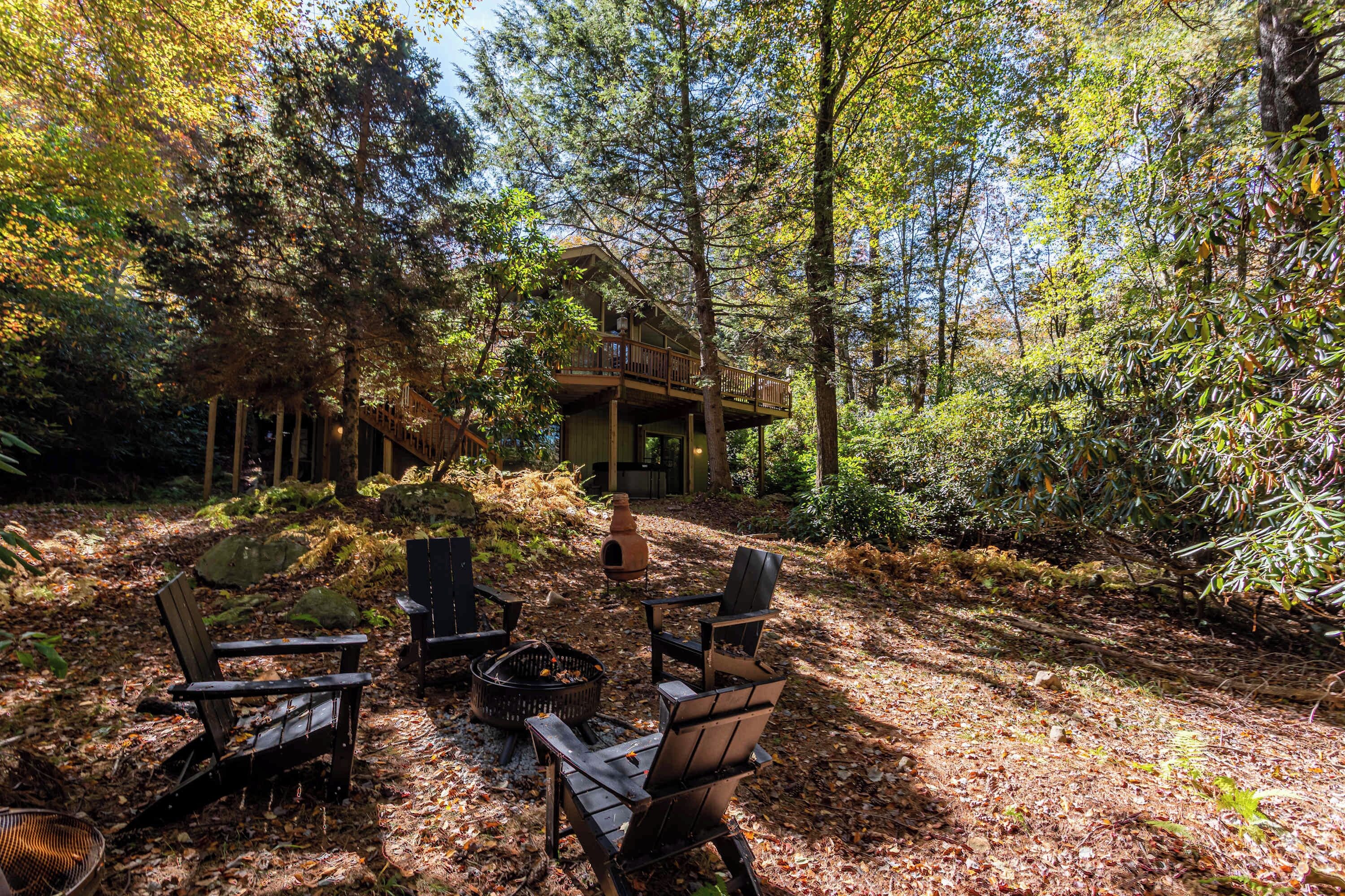 A cabin with a deck, surrounded by trees and a fire pit with chairs.