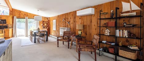 Een gezellige, goed verlichte kamer met houten wanden, een eethoek met stoelen en een tafel, een boekenkast met diverse spullen, en een airconditioner.