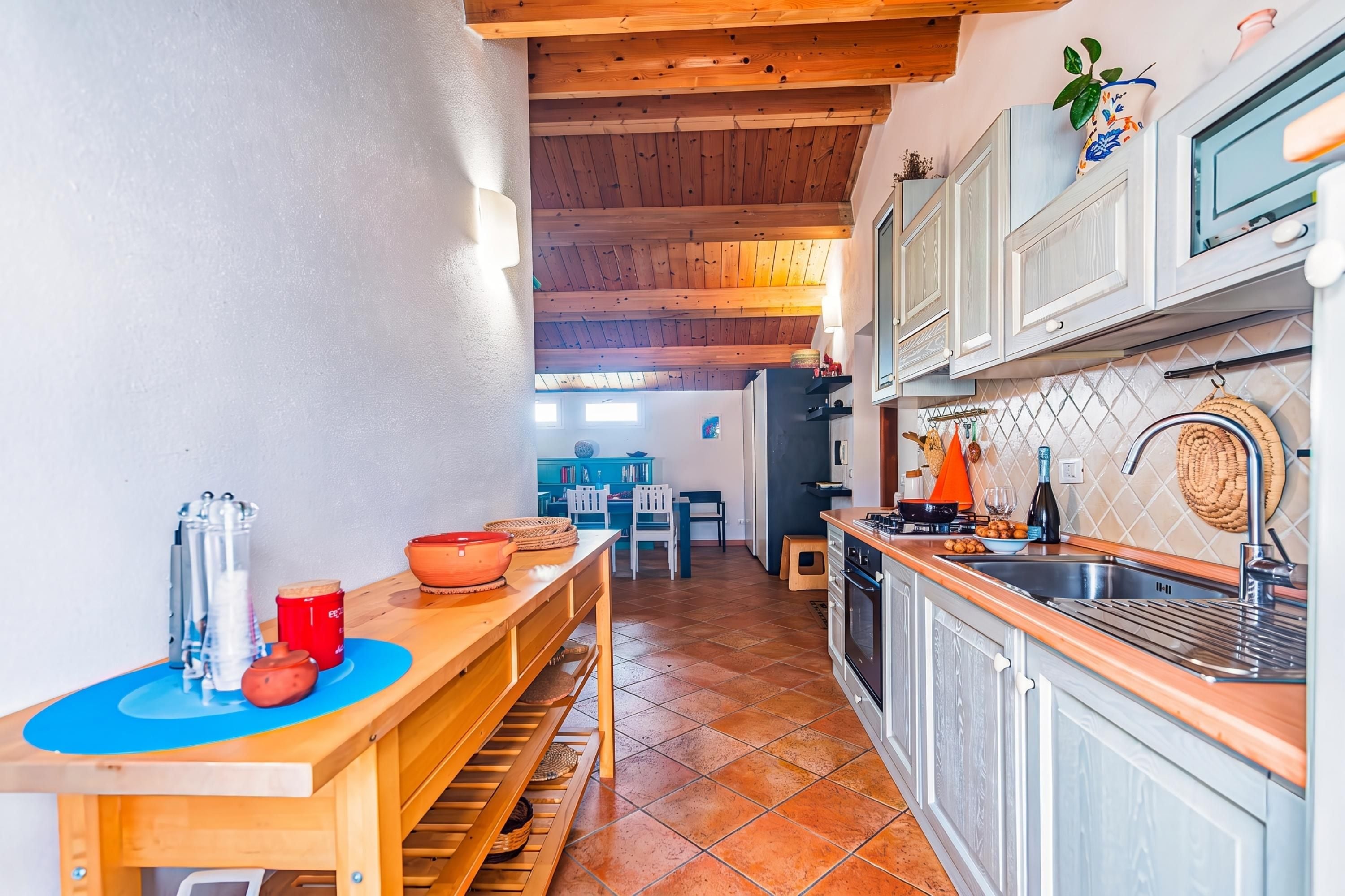 Cozinha com vigas de madeira, área de refeições com cadeiras e vista para um quarto azul ao fundo.