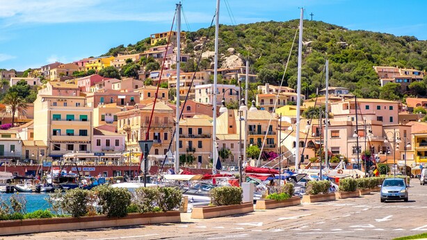 A coastal town with colorful buildings, a marina with boats, and a hill in the background.