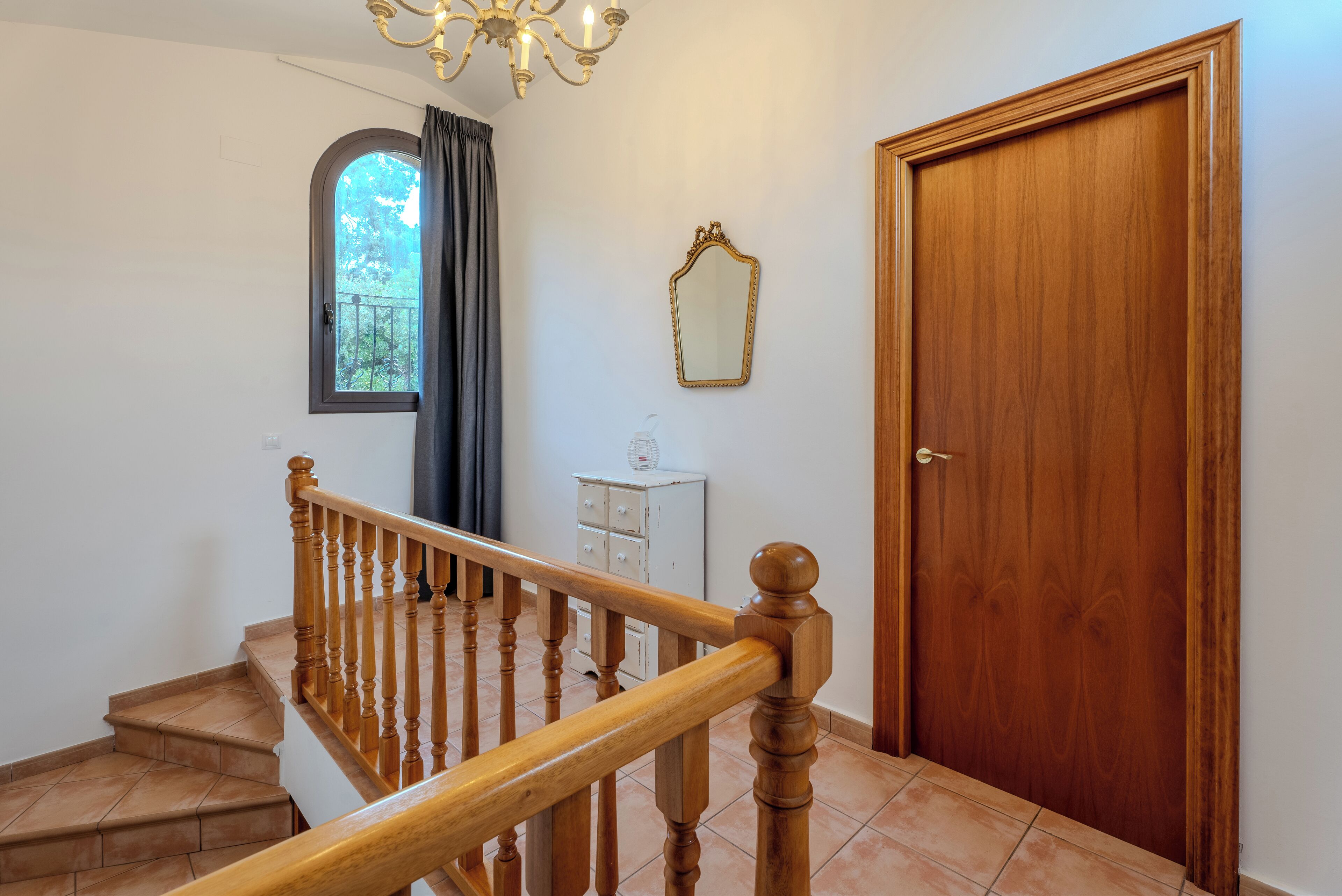 A staircase with wooden balusters, a chandelier, a wooden door, and a window with curtains.