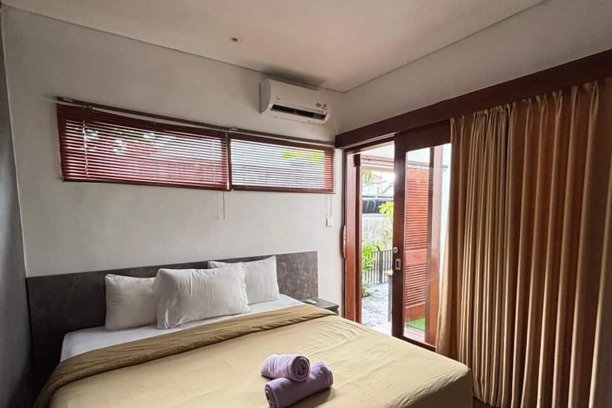 A hotel room with a large bed, a window with blinds, an air conditioning unit, and a view of the outside.