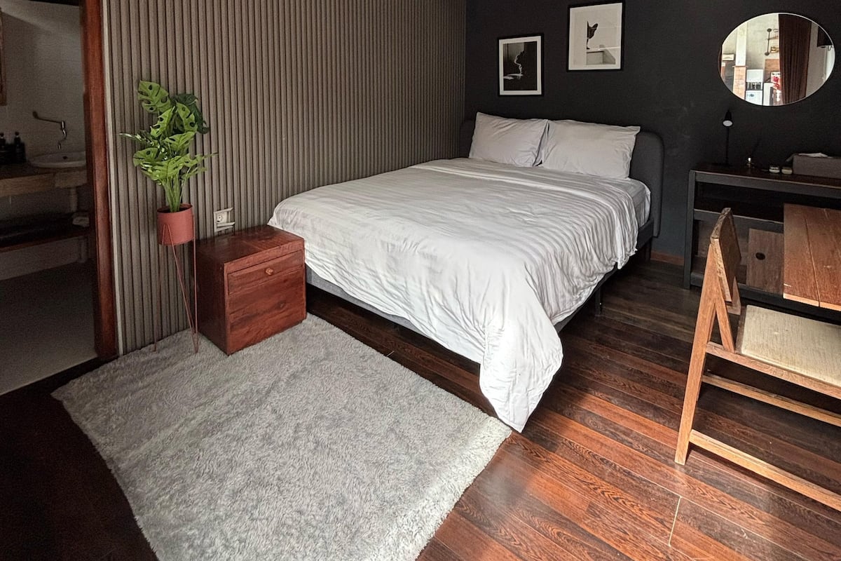 A modern bedroom with a large bed, a small desk, a potted plant, and a mirror on the wall.