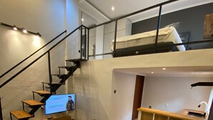 En moderne lejlighed i loftstil med trappe op til en soveplads i loftet, et lille køkken og et opholdsområde med sofa og skammel.
