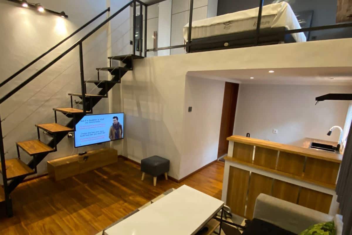 A modern loft-style apartment with a staircase leading to a loft bed, a small kitchen area, and a seating area with a sofa and ottoman.