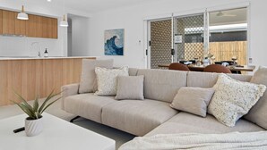 A modern living room with a beige sectional sofa, a white coffee table, and a potted plant. There is a kitchen area with wooden cabinets and a white countertop. A dining area with a table and chairs is visible through a glass door. - Secluded 4-Bed Retreat Mins to Beaches & Wineries (Victoria Point)