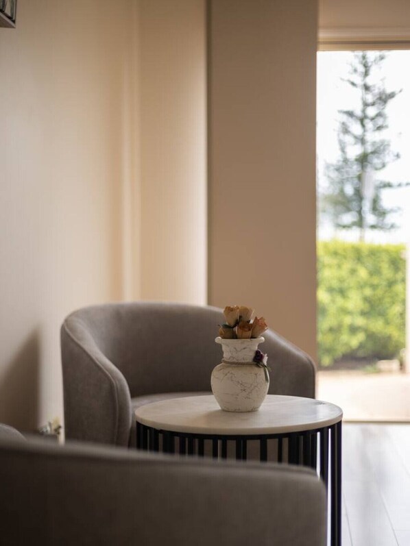 A room with a grey armchair, a small round table with a white vase containing dried flowers, and a view of greenery outside. - Ocean View From Balcony 5BR Home w/Pool @Redcliffe (Clontarf)