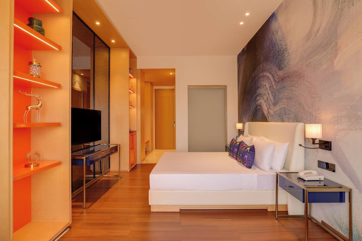 Ein modernes Hotelzimmer mit einem großen Bett, einem Fernseher, einem Schreibtisch mit Lampe und einem großen, abstrakten Gemälde an der Wand.