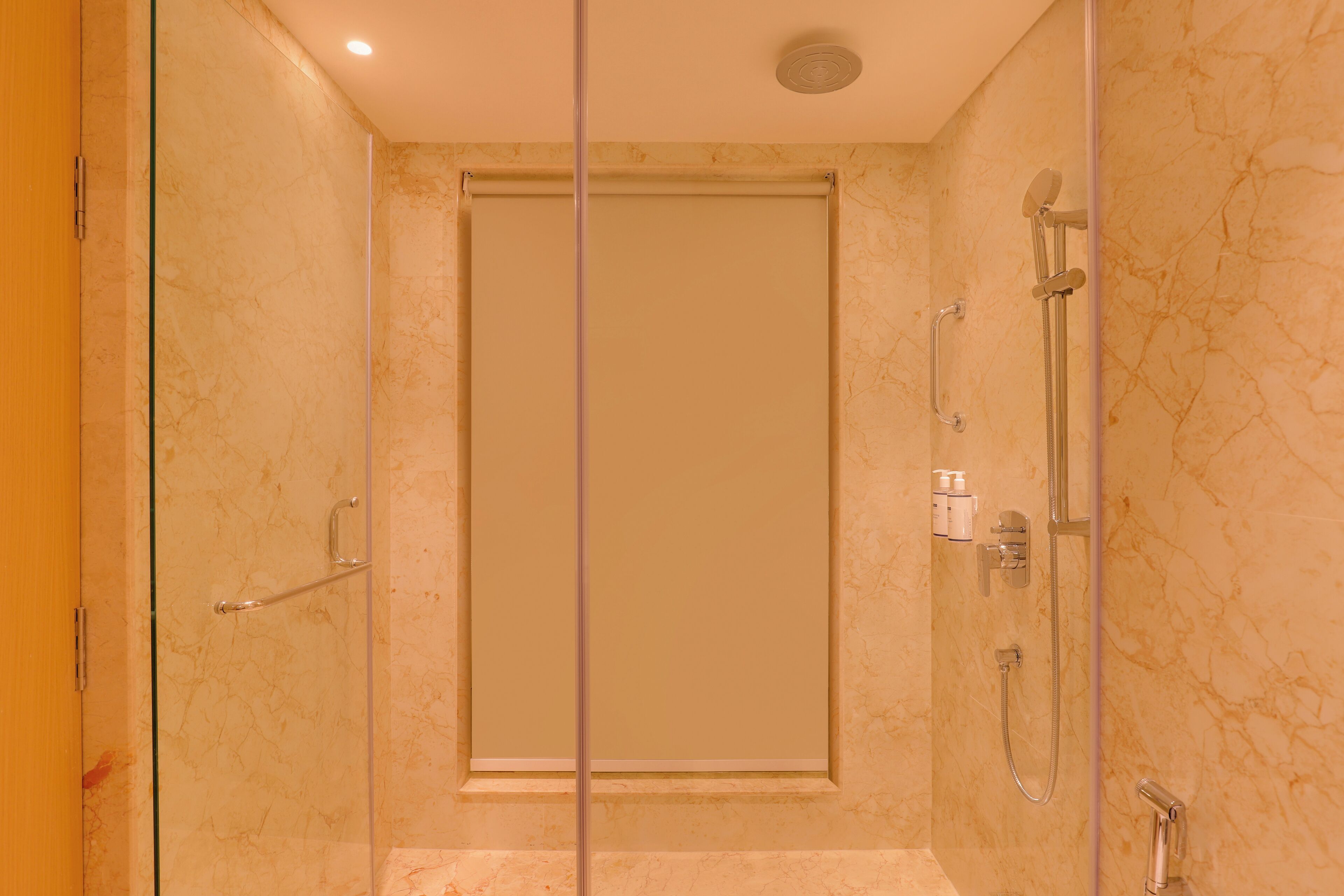 A modern bathroom with a glass-enclosed shower, marble walls, and a visible toilet paper dispenser.