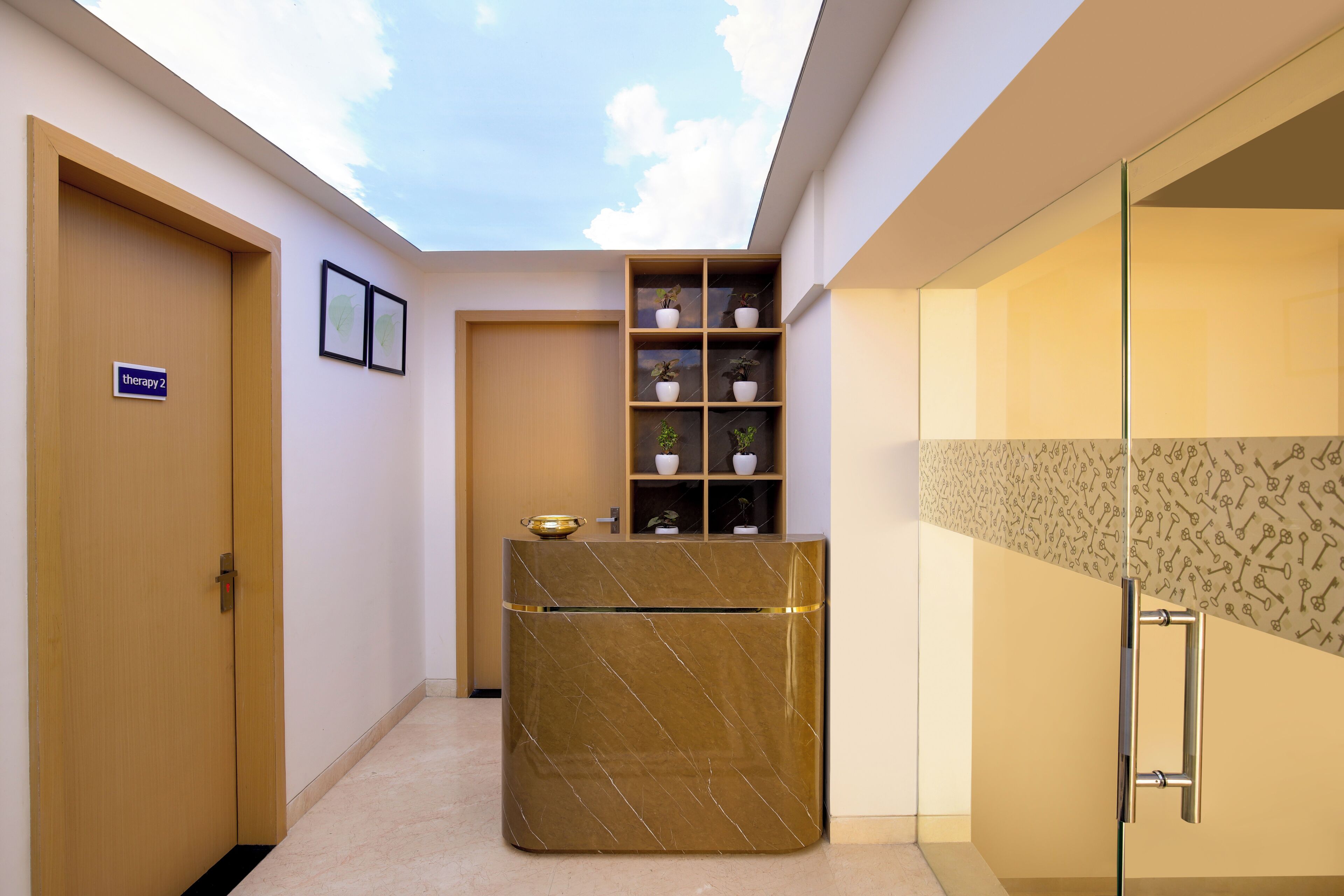 A modern hotel lobby with a reception desk, a sign for therapy room 2, and a glass partition with a patterned wall.