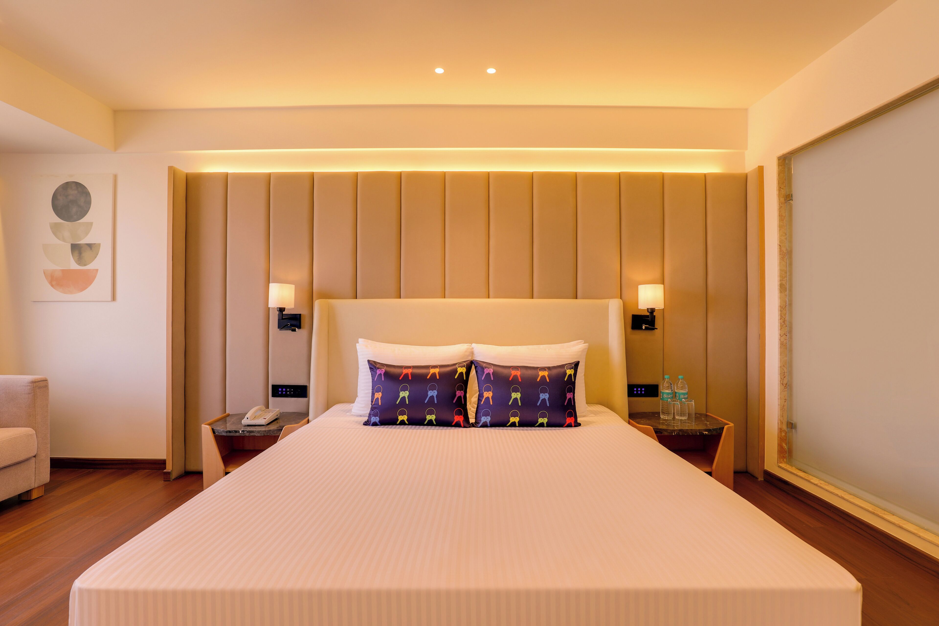 A hotel room with a large bed, two bedside lamps, a small table with a phone and water bottles, and a chair.