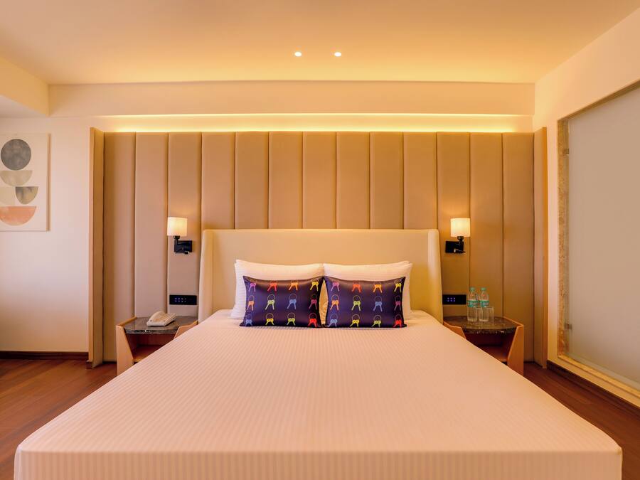 A hotel room with a large bed, two bedside lamps, a small table with a phone and water bottles, and a chair.