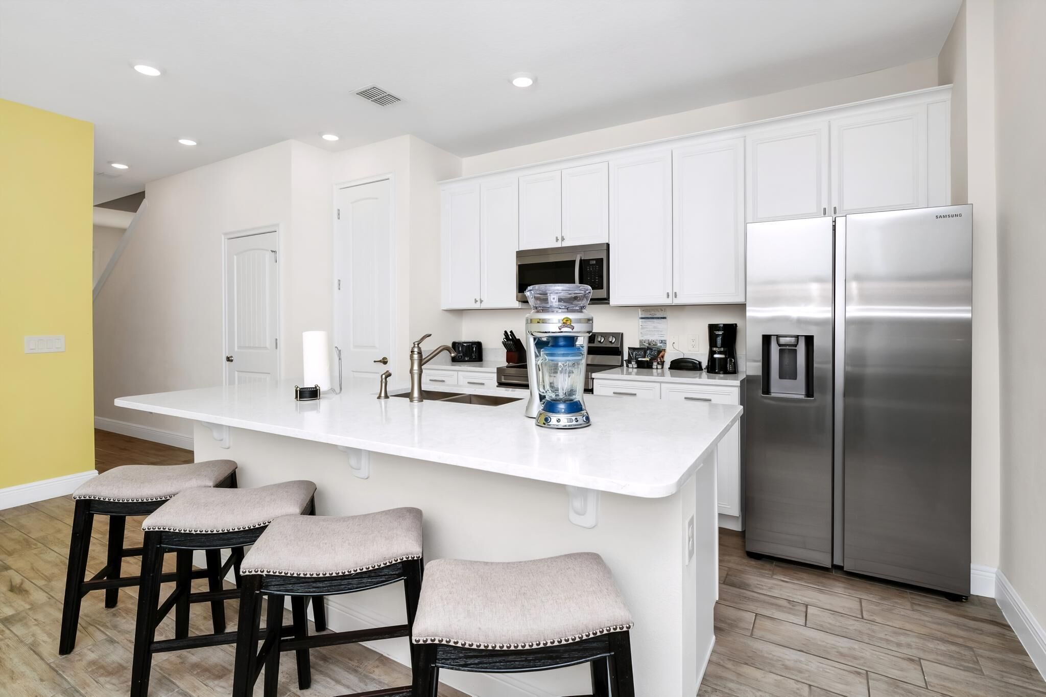 A modern kitchen with white cabinets, a stainless steel refrigerator, a blender, and bar stools.