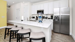 A modern kitchen with white cabinets, a stainless steel refrigerator, a blender, and bar stools.