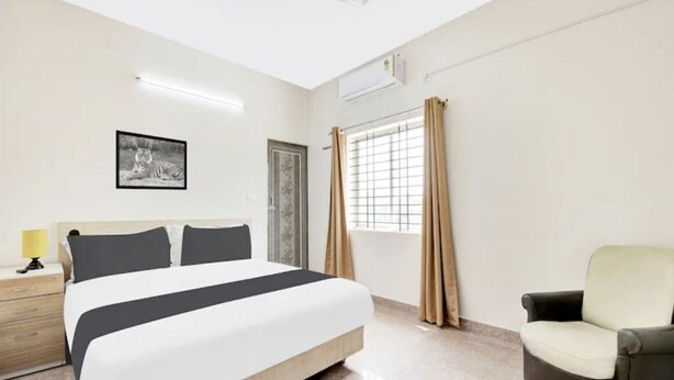 A hotel room with a bed, a chair, a nightstand, a window with curtains, and a wall-mounted air conditioner. - Welcome Inn Express (Bengaluru)