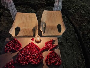 Two wicker chairs with a candle and red rose petals on a wooden surface. - Vivaans Beach Resort (Canacona)