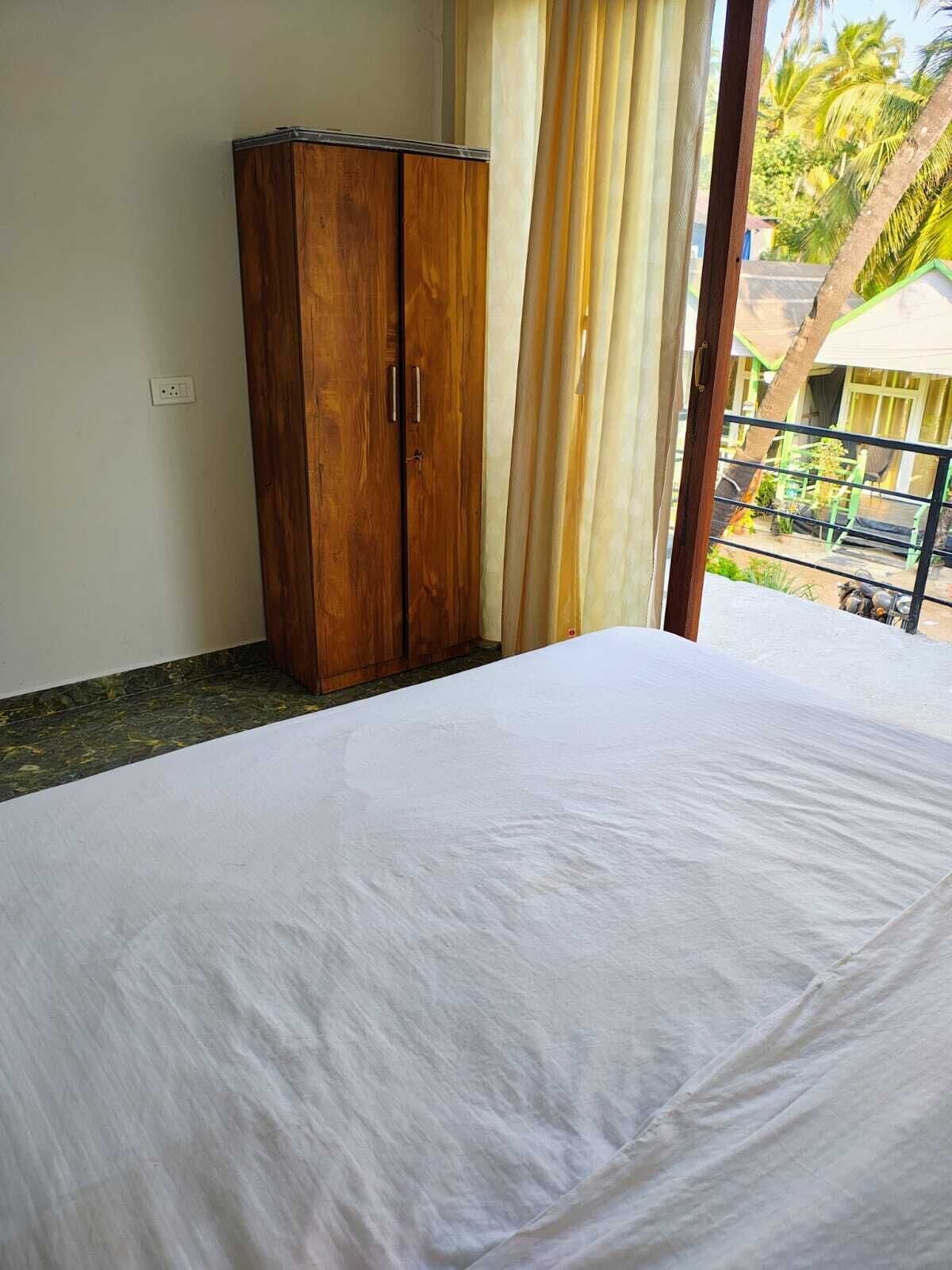 A room with a bed, wooden wardrobe, and a view of a balcony with palm trees.