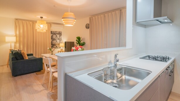 A modern kitchen with a central island, stainless steel sink, and a gas stove. There is a dining area with a white table and chairs, and a sofa in the background. - Seiwa Travel Time Sunahama (Osaka)