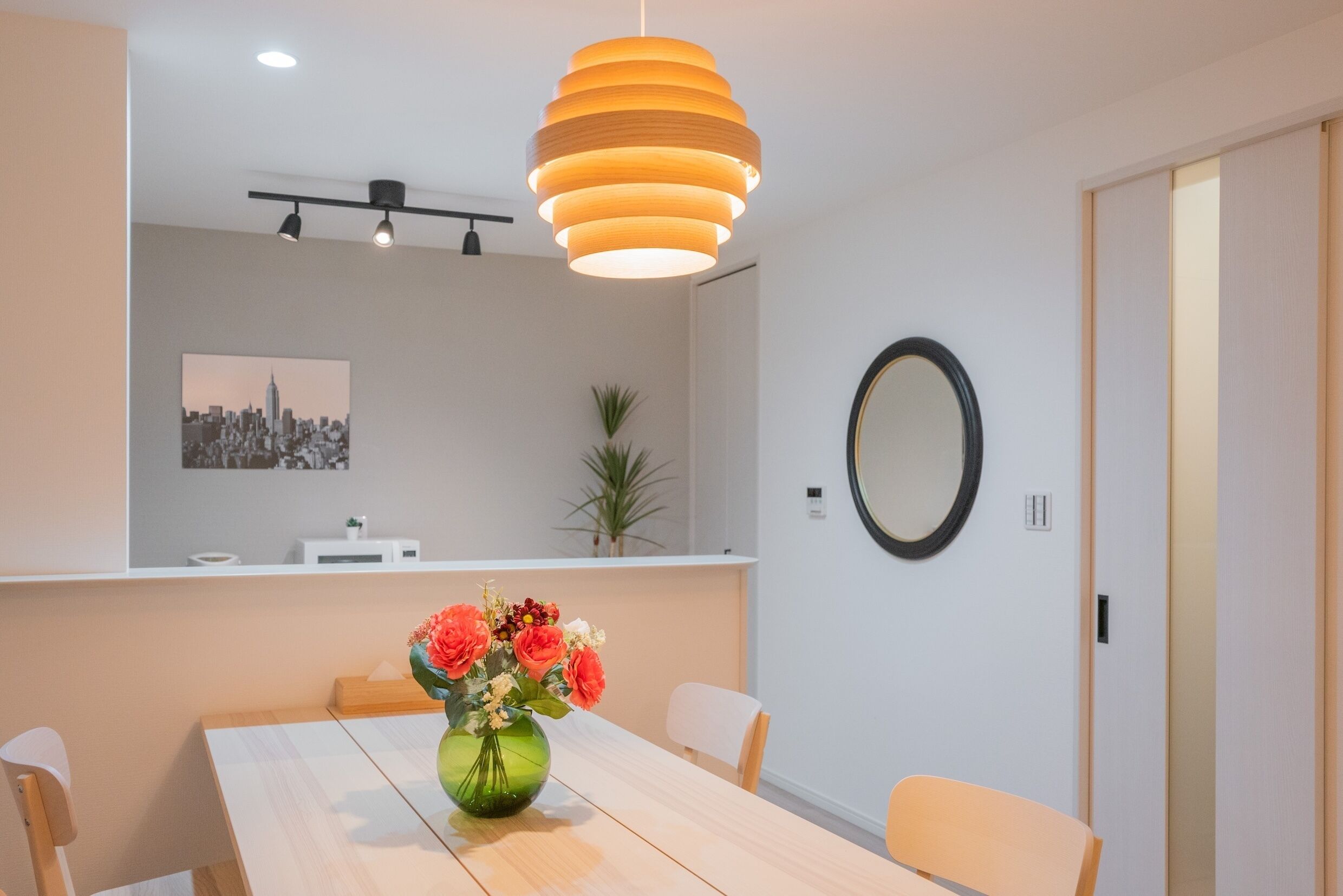 A modern dining area with a wooden table, chairs, a vase of flowers, a hanging light fixture, a cityscape painting, and a mirror on the wall.