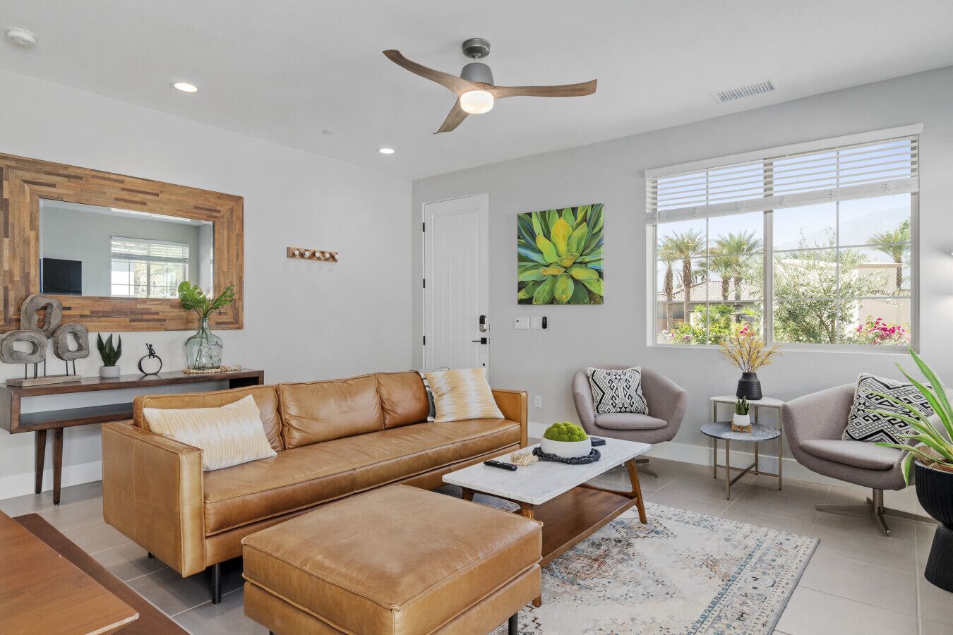 A modern living room with a brown leather sofa, a coffee table, and a grey armchair. There is a large window with blinds, a ceiling fan, and a wooden console table with decorative items.