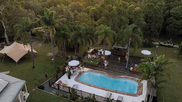 An outdoor pool area with a wooden deck, surrounded by palm trees and a mountainous backdrop.