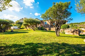 Uma villa de pedra com um jardim bem cuidado, árvores e um céu azul límpido.