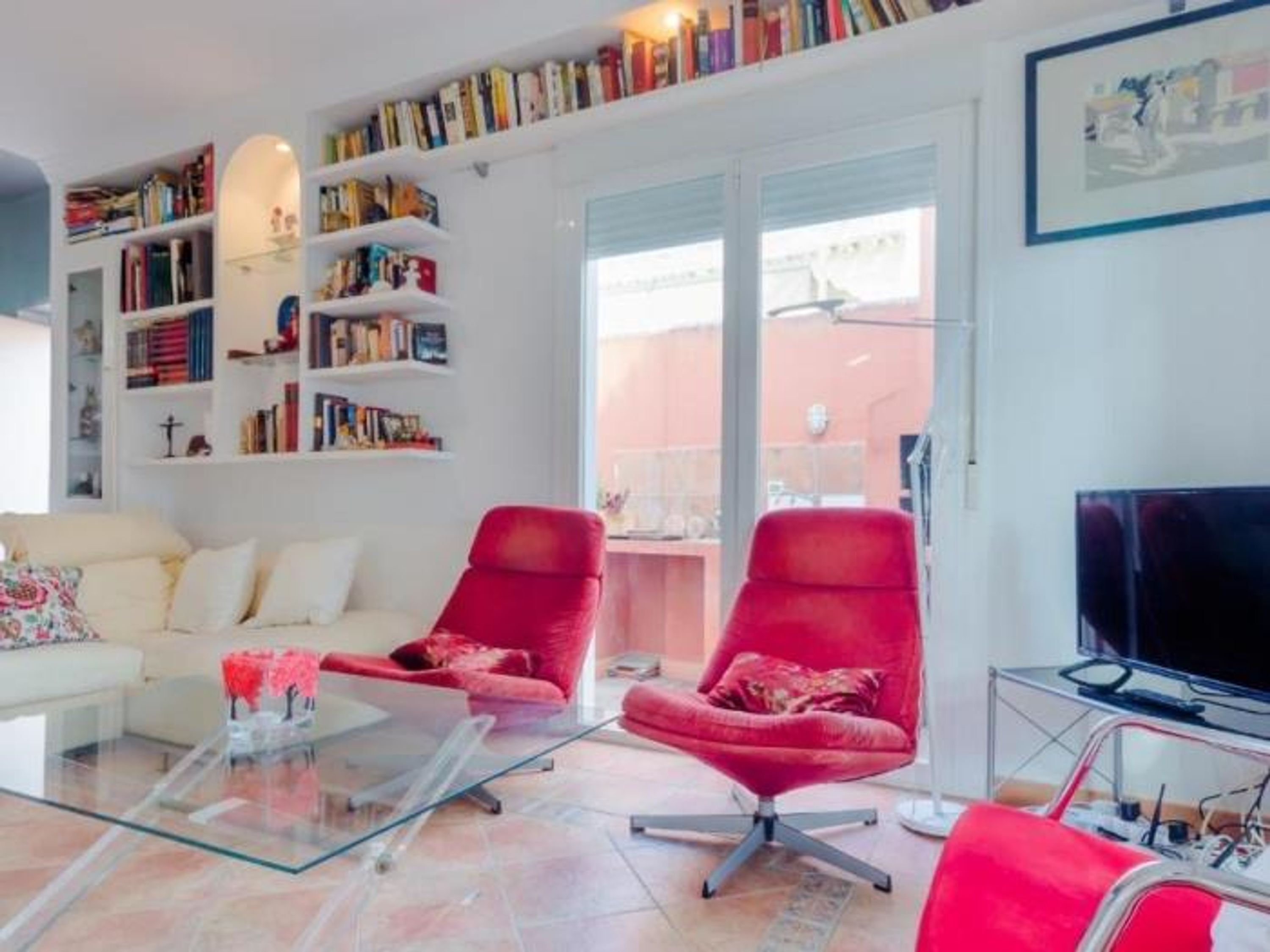 A modern living room with a white sofa, red chairs, a glass coffee table, and a bookshelf filled with books.