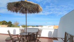Una terraza con mesa y sillas, un paraguas de paja y vistas al mar y edificios.
