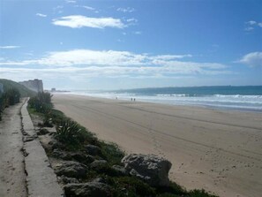 Una playa con un paseo de concreto, afloramientos rocosos y un edificio distante.