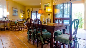A dining area with a wooden table and chairs, a clock on the wall, and a window with curtains. - Villa La Vega 14354 in Chiclana (Chiclana de la Frontera)