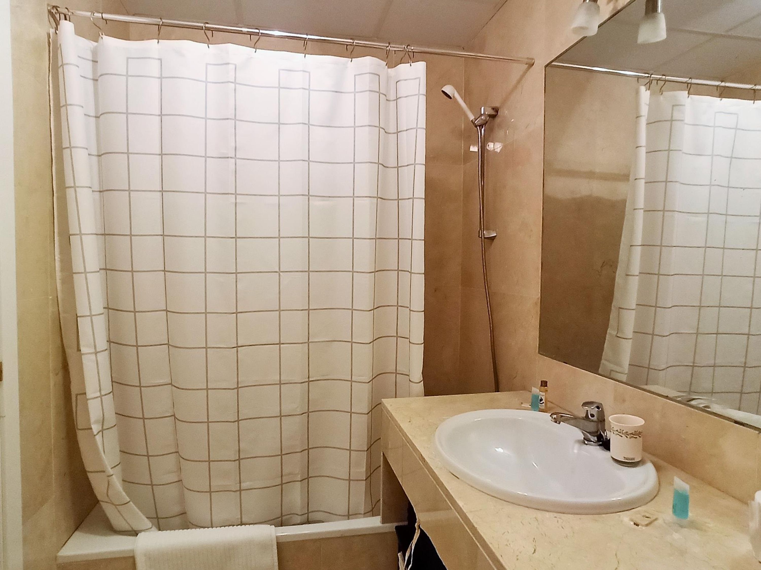 A bathroom with a grid-patterned shower curtain, a sink with a cup and a bar of soap, and a mirror reflecting the shower area.
