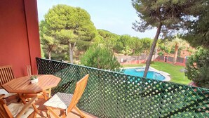 A balcony with a wooden table and chairs, a pool, and a view of a green landscape with trees and a building in the background. - Dunas II 2D 15436 in El Puerto de Santa Mar a (El Puerto de Santa Maria)