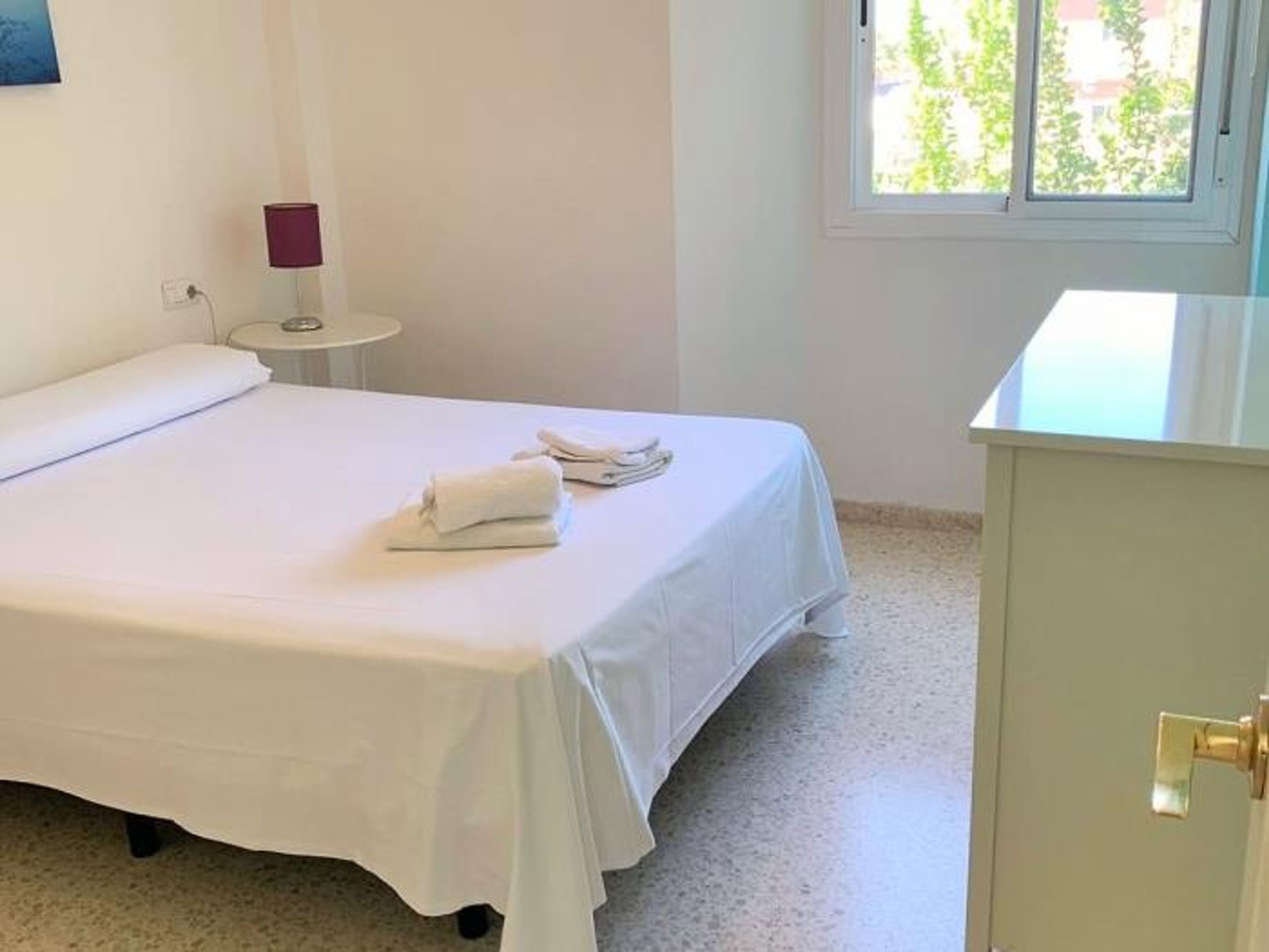 Habitación de hotel con cama, una mesita de noche con lámpara y ventana con vistas a zonas verdes.