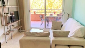 Una sala de estar moderna con un sofá beige, un televisor sobre un mueble bajo y un balcón con mesa y sillas.