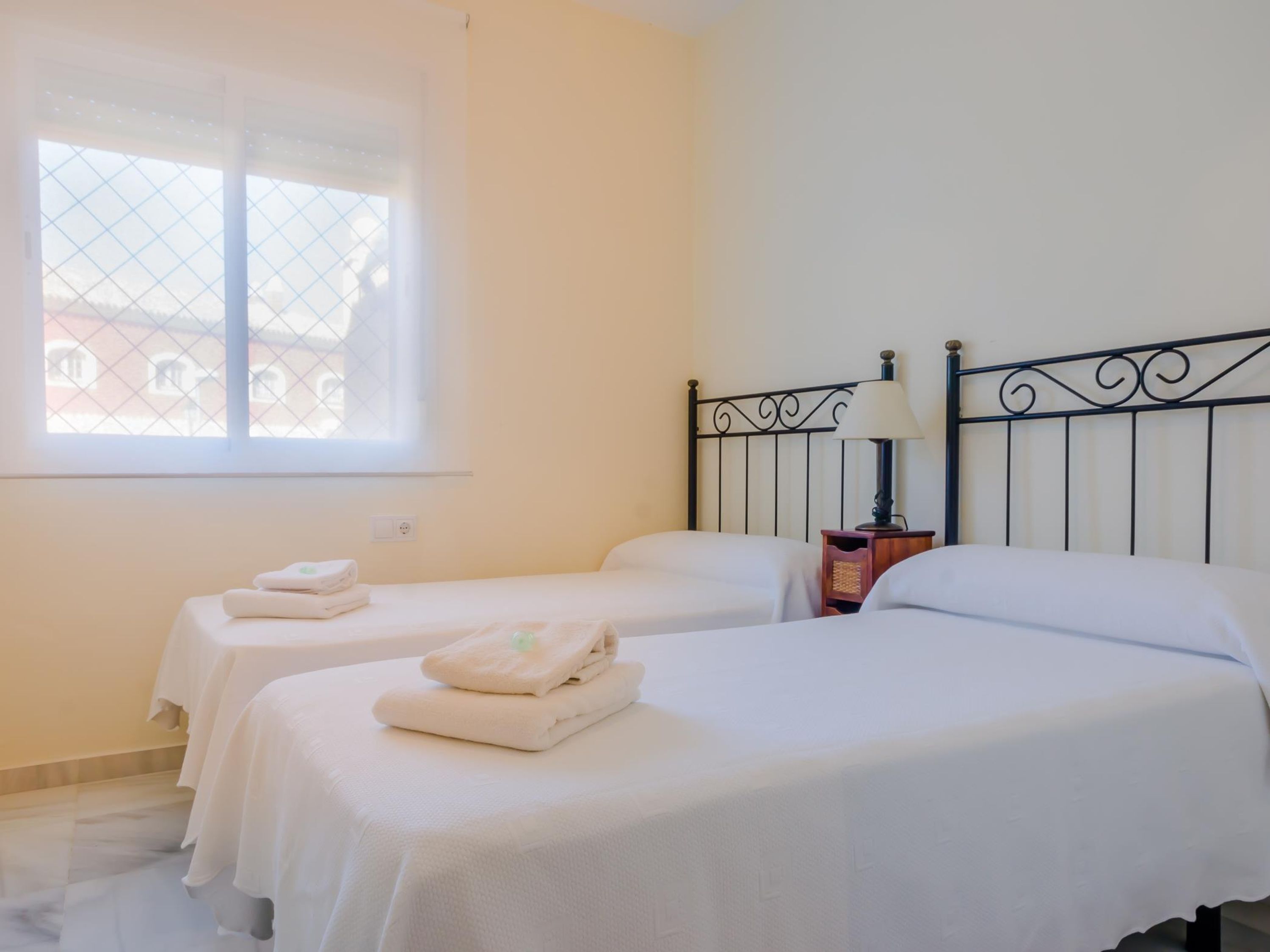A hotel room with two beds, a window with a view of buildings, and a bedside table with a lamp.